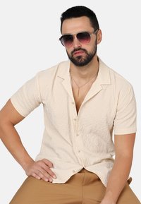 Kurzärmliges, hellbeiges Button-up-Hemd aus strukturiertem Stoff. Das Modell trägt Sonnenbrille in Dunkel und eine silberne Kettenhalskette, sitzt mit der Hand auf dem Oberschenkel.