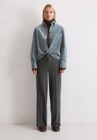 Lichtblauw overhemd met knopen, lange mouwen, borstzak, gedragen over een bruine coltrui. Grijze, wijde broek met een gestructureerde afwerking. Bruine schoenen.