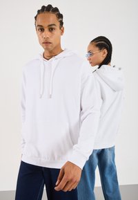 To personer med fletninger, iført hvide hættetrøjer og blå jeans, den ene vendt direkte imod kameraet, mens den anden står sidelæns mod en ensfarvet baggrund.