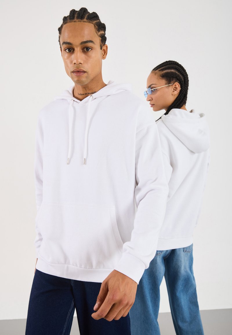 To personer med fletninger, iført hvide hættetrøjer og blå jeans, den ene vendt direkte imod kameraet, mens den anden står sidelæns mod en ensfarvet baggrund.