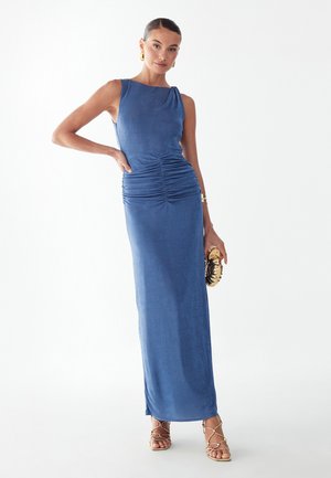 Mujer con vestido maxi azul sin mangas plisado, sosteniendo un bolso de mano dorado, con pendientes de aro dorados y sandalias de tiras, de pie con confianza.