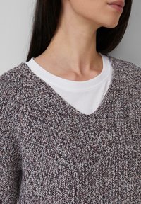 Frau mit langen dunklen Haaren, die einen mehrfarbigen strickwaren Pullover über einem weißen Rundhalsshirt trägt, zeigt ihren Hals und das untere Gesicht in neutraler Pose.