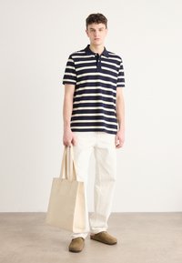 Stripet polo skjorte i marineblått og krem, i bomullsblanding, med krage og tre-knaps placket. Kombinert med beige lærveske og hvite jeans.