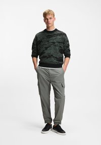 Νέος άνδρας όρθιος με τα χέρια στις τσέπες, φορώντας μαύρο και πράσινο πουλόβερ με σχέδια, γκρι παντελόνια cargo και μαύρα αθλητικά σε λευκό φόντο.