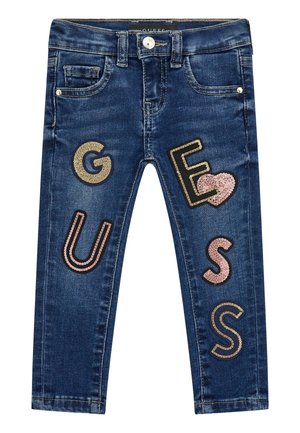 Jean en denim bleu foncé pour enfants avec des lettres en sequins formant le mot « GUESS » et un cœur rose en sequins sur la jambe droite.