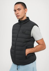 Crni prešani puffer prsluk s visokim ovratnikom, prednjim zatvaračem i bočnim džepovima. Uparen s sivom majicom, prikazan na neutralnoj pozadini.