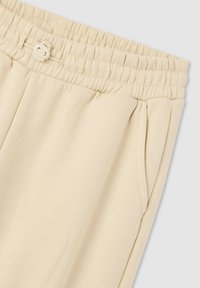 Beżowe dresy z elastycznym pasem, zapięciem na guzik, kieszeniami z przodu i gładką fakturą materiału. Posiadają pliski w pobliżu kieszeni.