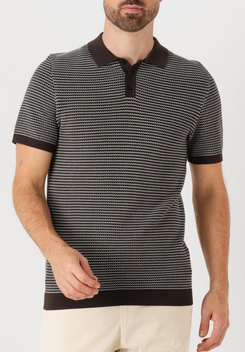 Homme portant un polo à manches courtes noir et blanc à motifs, avec col et poignets noirs, assorti à un pantalon beige clair.
