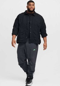Camicia nera trapuntata sopra una maglietta nera, abbinata a pantaloni grigio scuro con il logo verde della Nike. Sneakers nere con dettagli testurizzati.