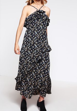 Femme portant une robe maxi noire sans manches avec un petit imprimé floral et des détails à volants, associée à des bottines noires.