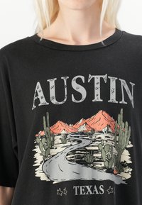 Juodas oversized t-shirt marškinėliai su užrašu "Austin Texas" ir grafiką vaizduojančia vingiuotą kelią su kalnais ir kaktusais švelnių spalvų tonais.