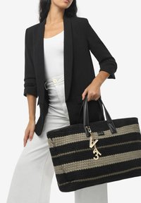 Bolso tote tejido de rayas negras y beiges con asas de cuero negro y un charm dorado. El bolso presenta una forma rectangular amplia y una tela texturizada.