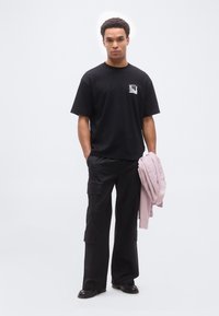 Άνδρας που φοράει μαύρο Τ-shirt και παντελόνι cargo, κρατώντας ένα ανοιχτό ροζ πουκάμισο, στέκεται μπροστά σε απλό λευκό φόντο.