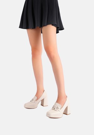 Scarpe con tacco a blocco beige, con finitura in pelle liscia, dotate di punta arrotondata e un dettaglio decorativo a catena sulla parte anteriore.