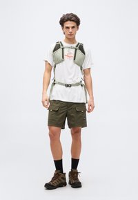 Νεαρός άνδρας που φοράει λευκό μπλουζάκι με σχέδιο, χακί cargo σορτς, ορειβατικά μποτάκια και χακί στηθικό ζώνη με λουριά και αγκράφες.