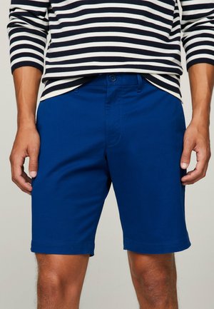 Mann trägt blaue knielange Shorts und ein schwarz-weiß gestreiftes Langarmshirt, steht mit entspannten Händen an den Seiten.