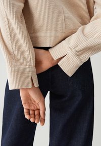 Beige Cordjacke mit geripptem Textur, die eine taillierte Passform und cinchbare Manschetten aufweist, kombiniert mit dunkelblauen Jeans. Hand hinter dem Rücken positioniert.