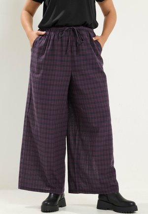 Persona que lleva pantalones anchos de cuadros púrpura y negro con bolsillos delanteros y botas negras hasta el tobillo, manos en los bolsillos.