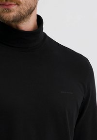 Mann in schwarz, der einen Pierre Cardin Rollkragenpullover trägt, Nahaufnahme des Halses und des oberen Körpers vor einem einfarbigen Hintergrund.