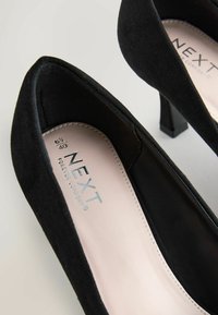 Chaussures à talons hauts en daim noir avec un design à bout pointu, dotées d'un petit talon bloc et d'une semelle intérieure beige avec un branding embossé.