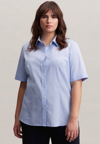 Camicia azzurro chiaro a maniche corte in cotone, con apertura a bottoni, colletto a punta e orlo leggermente curvato.
