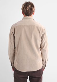 Camisa de pana beige con cuello abotonado, mangas largas y dobladillo redondeado. Presenta una textura de costillas verticales y una pequeña etiqueta de la marca en la parte trasera.