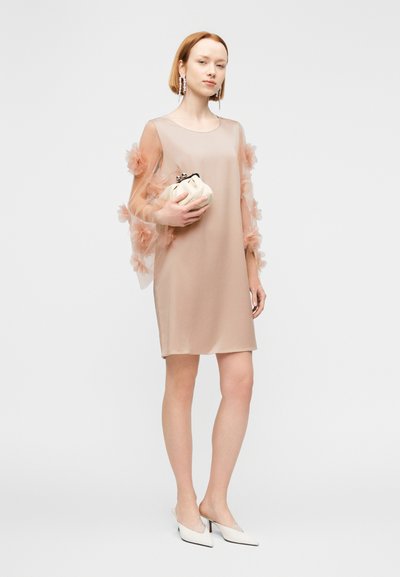 Γυναίκα που φοράει ένα διάφανο μπεζ φόρεμα με φλοράλ μανίκια, κρατώντας μια κρεμ τσάντα, φορώντας λευκές μυτερές γόβες και κρεμαστά σκουλαρίκια, στέκεται μπροστά σε λευκό φόντο.