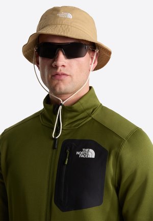 Man draagt een beige bucket hat, zwarte zonnebril en een olijfgroene ritssluiting jas met een zwart borstzakje en The North Face-logo's.