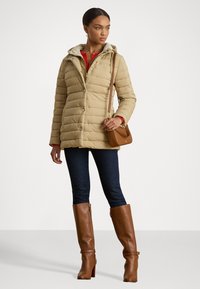 Casaco puffer bege com capuz com fecho e botões de pressão, combinado com jeans de ganga e botas de salto altas castanhas até ao joelho. Design e textura simples.