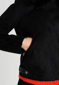 Czarna kurtka z fałszywej zamszowej skóry z zippowanym kieszeniem, czarnymi skórzanymi akcentami i żywym czerwonym dzianinowym wykończeniem. Kołnierz jest puszysty i teksturowany.