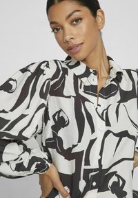 Blusa blanca con un audaz patrón abstracto negro, con mangas abullonadas, un cuello y cierre frontal de botones. Textura de tela suave y ligera.