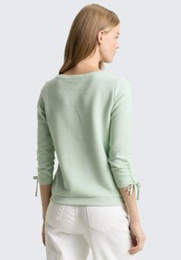 Lichtgroene gebreide trui met drie-kwart mouwen, voorzien van geplooide manchetten die zijn vastgebonden met strikjes. De achterkant heeft een brede halslijn en een geribde zoom.