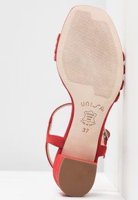 Sandale à talons hauts en daim rouge avec une semelle beige texturée, une bride à la cheville et un détail de boucle latérale. Présente des marquages en relief de la marque et de la taille.