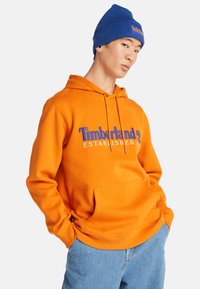 Felpa arancione con cappuccio e tasca frontale, caratterizzata dal logo blu con la scritta "Timberland". Indossata con un berretto di lana blu e jeans chiari.