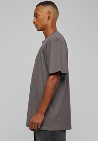 Un homme de profil portant un t-shirt gris à manches courtes, ample, et un pantalon foncé, contre un fond clair uni.