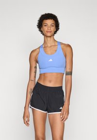 Lyseblå sportsbh med scoop-hals og fremre sømmer. Svarte shorts med hvit kant og elastisk midjebånd. Adidas-logoen er synlig.