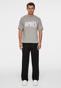 Grå bomuld T-shirt med fed hvid tekst "APRÈS," kombineret med sorte bukser og hvide sneakers. Simpelt, afslappet design.