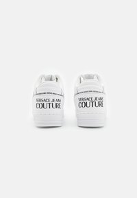 White leather sneakers with textured accents, featuring a flat sole and embossed "VERSACE JEANS COUTURE" logo on the heel.