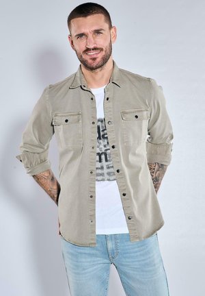 Helles graues Button-up-Hemd mit zwei vorderen Taschen, hochgekrempelten Ärmeln, kombiniert mit einem weißen Graphic T-Shirt und hellblauen Jeans.