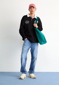 Mees roosa mütsi, musta pika varrukaga särgi, siniste teksade ja spordijalatsitega, hoiab rohelise kandekoti taustal valge seina ees.