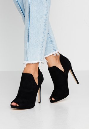 Bottines noires en daim à talons aiguilles avec des découpes sur les côtés et un bout ouvert, portées avec des jeans bleu clair à ourlet effiloché.