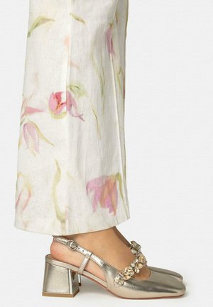 Glänzende goldene Blockabsätze mit großen Schmucksteinverzierungen, getragen zu weißen Hosen mit zartrosa und grünen Blumenmustern.