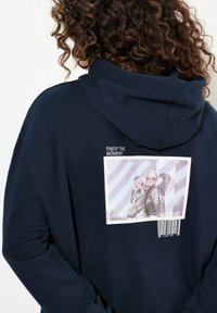 Felpa blu navy con tasca frontale, caratterizzata da una stampa grafica rettangolare con il testo "GODITI IL MOMENTO" e un codice a barre. Texture morbida.