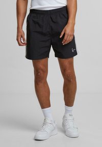Shorts noirs avec taille élastique, présentant des rayures sur les côtés et un logo blanc. Portez-les avec des baskets blanches et des chaussettes ; tissu léger et texturé.
