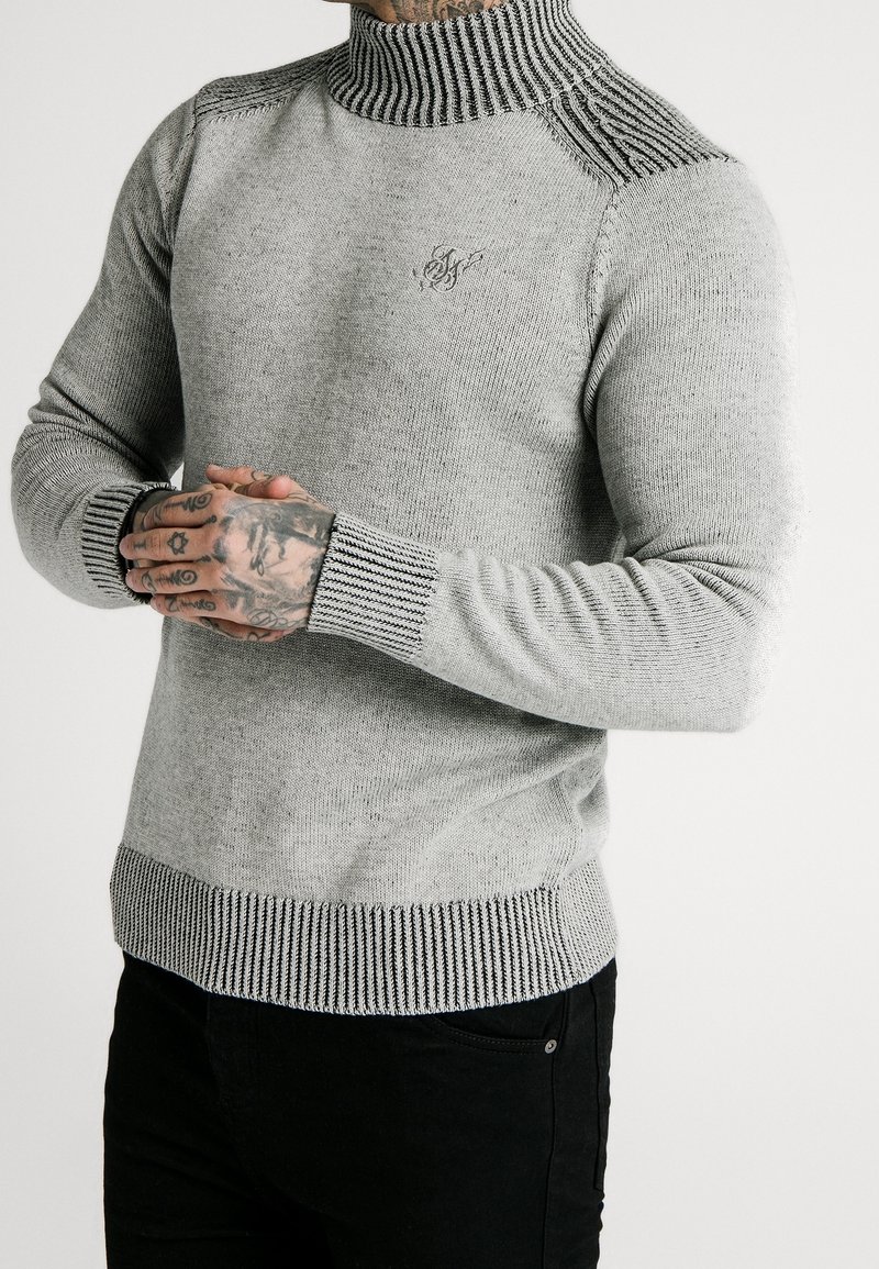 Homme portant un pull à col roulé en maille gris clair avec des poignets, un col et un ourlet côtelés noirs et blancs, associé à un pantalon noir, les mains croisées.