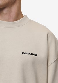 Felpa beige con scollatura rotonda e bordi a costine. Presenta un ricamo nero "PEGADOR" sul petto. Texture morbida e vestibilità comoda.
