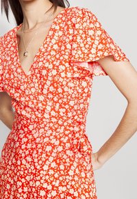 Červené květinové šaty s krátkými volánkovými rukávy a výstřihem do V. Mají zavinovací design s bílými a růžovými květinovými vzory.