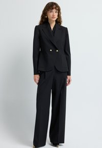 Zwarte op maat gemaakte pak met een dubbelzijdige blazer met gouden knopen, wijde broekspijpen en een soepele stofstructuur.