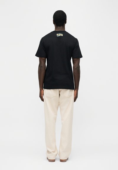 Άνδρας στέκεται με την πλάτη, φοράει μαύρο T-shirt με την επιγραφή «Billionaire Boys Club» στην πάνω πλάτη και μπεζ παντελόνι.