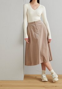 Vrouw leunend tegen een muur, gekleed in een crèmekleurig geribbeld shirt met lange mouwen, een beige corduroy midi-jurk, crèmekleurige sokken en beige sneakers.